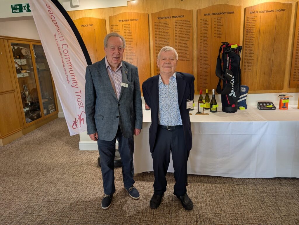 Tony, Chair of Dacorum Community Trust (left), and Geoff Budd, longstanding trustee (right), standing together in front of the raffle prize table and a Dacorum Community Trust branded banner in the clubhouse at Berkhamsted Golf Club's Seniors Golf Day. Behind them, bottles of wine, a Srixon golf bag, and other prizes are visible, alongside the club's historic honours boards.