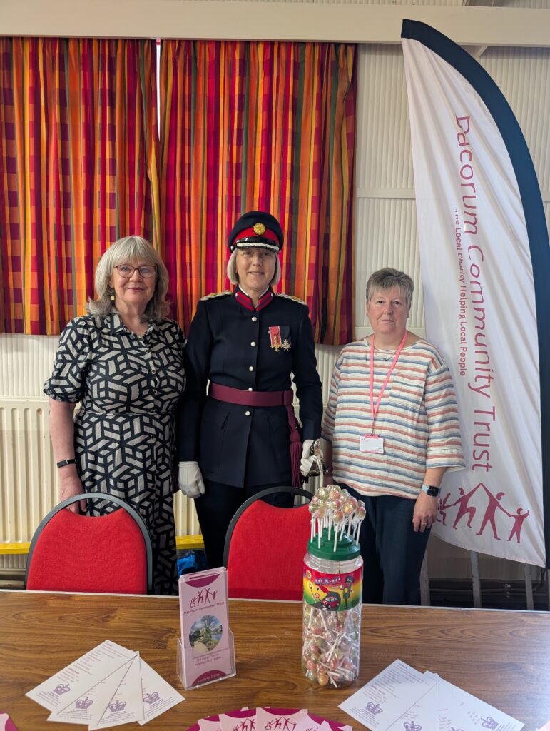 Three women stand together smiling in front of a Dacorum Community Trust banner. On the left is the Trust Manager in a patterned dress, in the centre is Deputy Lieutenant of Hertfordshire Liz Green OBE in full ceremonial uniform, and on the right is volunteer Sarah in a striped top wearing a lanyard. A table in the foreground displays Dacorum Community Trust leaflets and a jar of lollipops. The event was held at the Kings Langley Local Charity Event during Volunteers Week 2026.