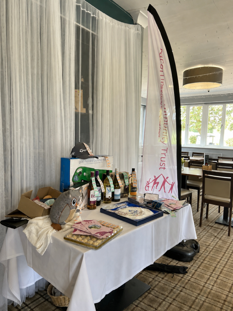 A table set with a white cloth displaying a range of raffle prizes, including bottles of wine and spirits, chocolates, a decorative owl ornament, a golf cap, and other gifts, alongside a Dacorum Community Trust branded banner, in the function room at Redbourn Golf Club.