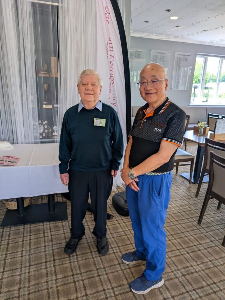 ony, Chair of Dacorum Community Trust, and Philip, trustee of Dacorum Community Trust and member of Redbourn Golf Club, standing together inside the club's function room in front of a Dacorum Community Trust branded banner at the Senior Charity Golf Day.
