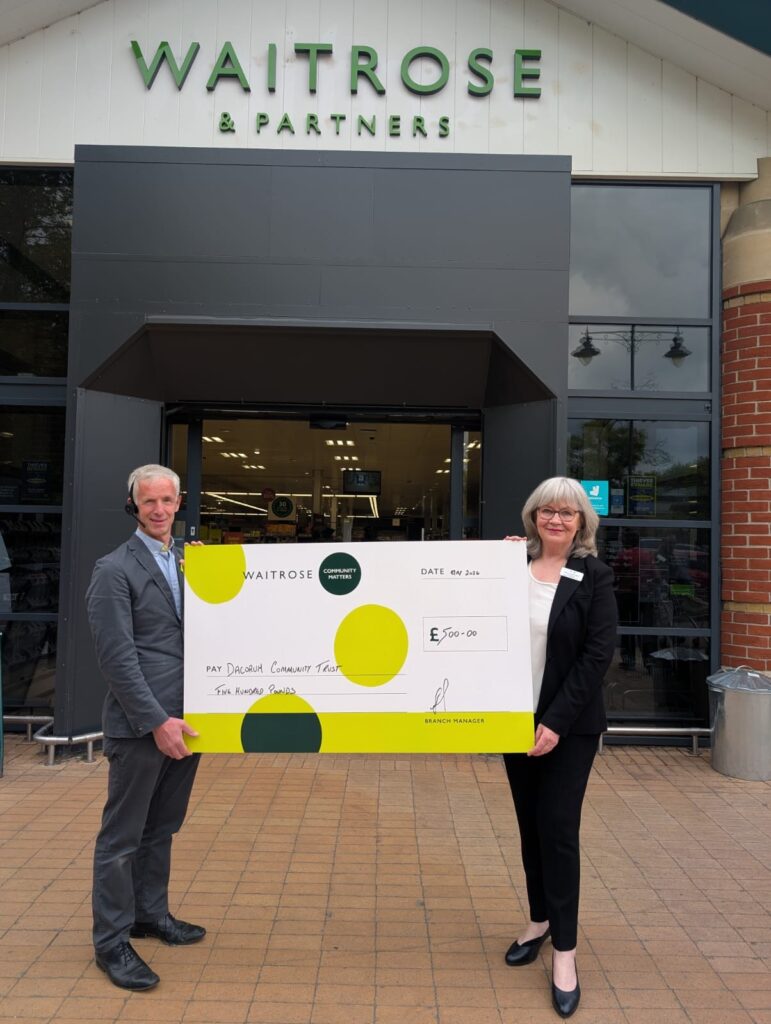 Dena, Trust Manager of Dacorum Community Trust, and a member of the Waitrose Berkhamsted team stand outside the store entrance holding a large ceremonial cheque made out to Dacorum Community Trust for five hundred pounds, presented through the Waitrose Community Matters scheme.
